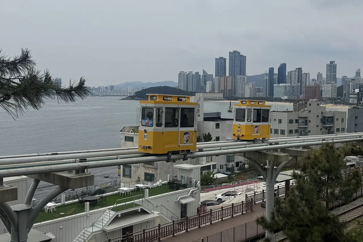 Two yellow Sky Capsule cabins on the elevated track above the Busan shore, with the Haeundae skyline across the bay
